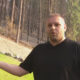 Man in a black T-shirt speaks outdoors with his left arm extended, standing on grass with pine trees and a fence in the background.