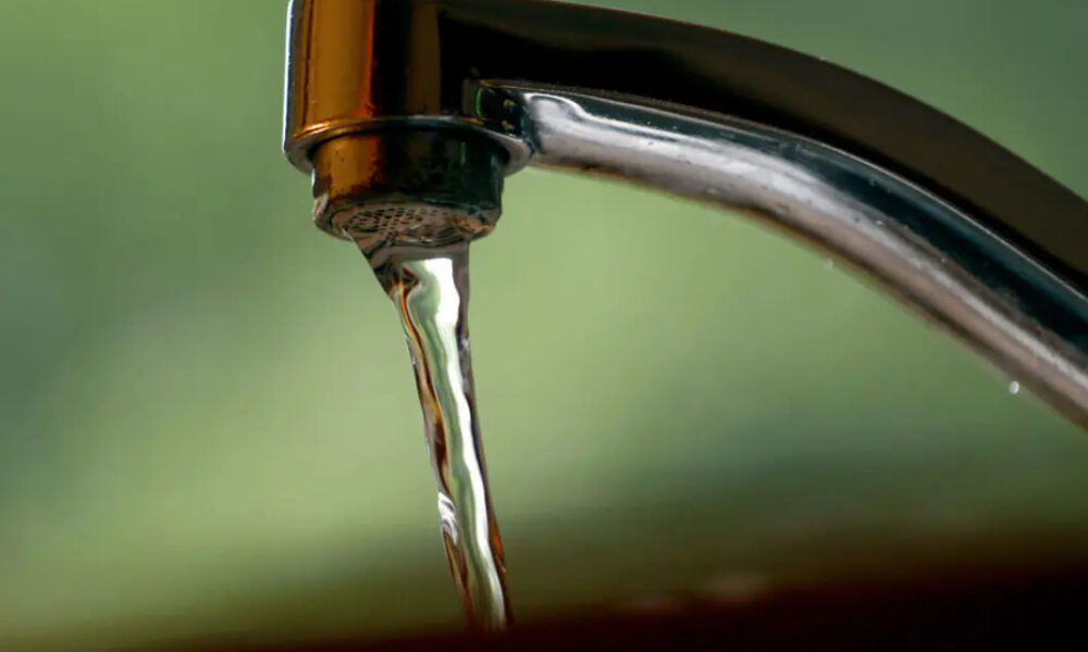 Close-up of a chrome faucet with a single stream of water dripping into a sink, blurred green background behind it.