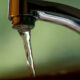 Close-up of a chrome faucet with a single stream of water dripping into a sink, blurred green background behind it.