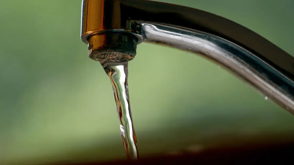 Close-up of a chrome faucet with a single stream of water dripping into a sink, blurred green background behind it.