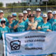 Youth baseball team in teal jerseys posing with banner that reads 'Washington Little League State Champions 2026' on a sunny field.