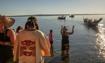 People in colorful festival attire stand in shallow water by the shore while small boats sail in the calm river ahead under a sunny sky.