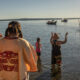 People in colorful festival attire stand in shallow water by the shore while small boats sail in the calm river ahead under a sunny sky.