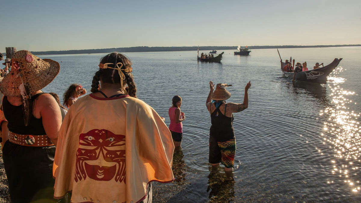 People in colorful festival attire stand in shallow water by the shore while small boats sail in the calm river ahead under a sunny sky.