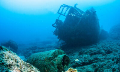 Underwater shipwreck resting on rocky seabed with blue ocean surrounding and small fish swimming nearby