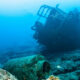 Underwater shipwreck resting on rocky seabed with blue ocean surrounding and small fish swimming nearby