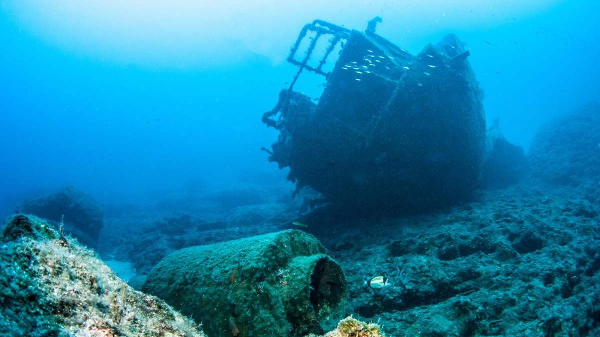Underwater shipwreck resting on rocky seabed with blue ocean surrounding and small fish swimming nearby
