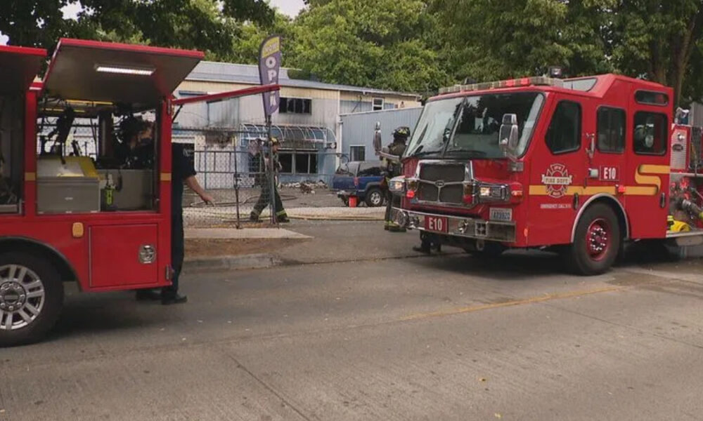 Two red fire trucks parked on a street with firefighters in gear near a chain-link fence and a building in the background.