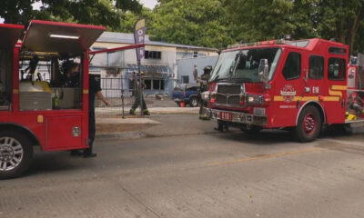 Two red fire trucks parked on a street with firefighters in gear near a chain-link fence and a building in the background.