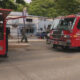 Two red fire trucks parked on a street with firefighters in gear near a chain-link fence and a building in the background.