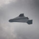 White unmanned aerial vehicle (drone) with swept wings flying through a cloudy sky.