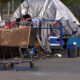 A cluttered roadside encampment with tents, a shopping cart, and scattered belongings beside a chain-link fence and a car.