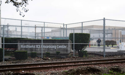 Chain-link fence surrounds an industrial area with a sign reading 'Northwest ICE Processing Center' and railroad tracks in the foreground.