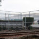 Chain-link fence surrounds an industrial area with a sign reading 'Northwest ICE Processing Center' and railroad tracks in the foreground.