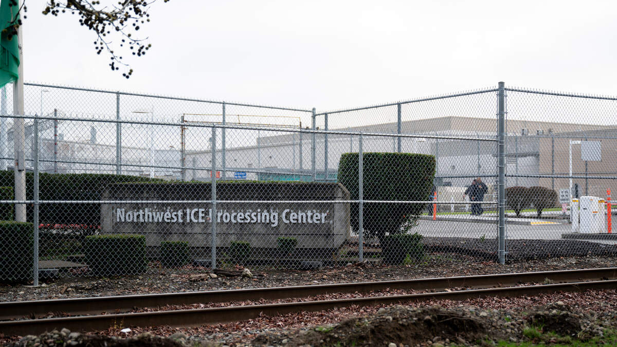 Chain-link fence surrounds an industrial area with a sign reading 'Northwest ICE Processing Center' and railroad tracks in the foreground.