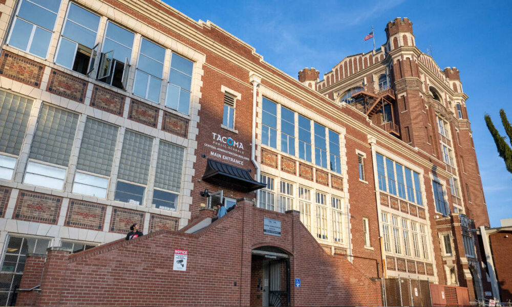 Brick, multi-story building with a tall tower on the right, large windows, and brick stairs leading to a main entrance under a small canopy. A U.S. flag waves atop the tower.