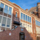 Brick, multi-story building with a tall tower on the right, large windows, and brick stairs leading to a main entrance under a small canopy. A U.S. flag waves atop the tower.