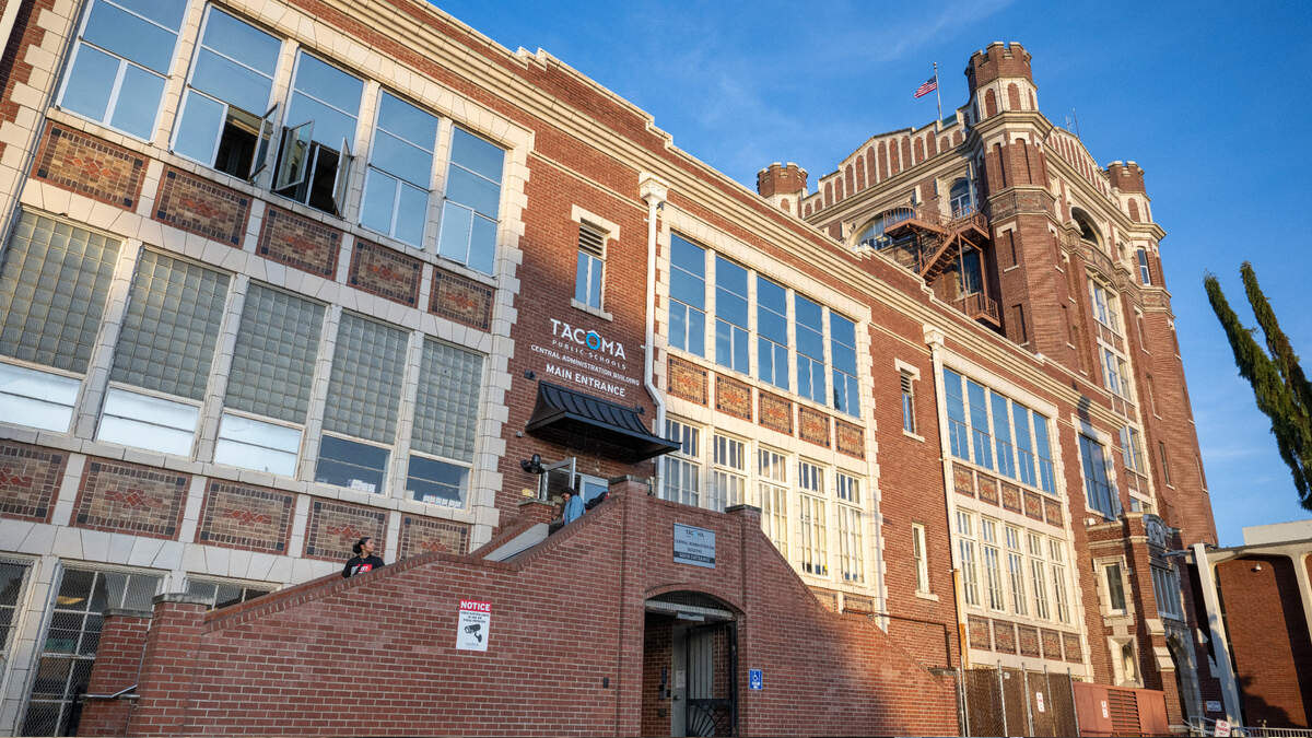 Brick, multi-story building with a tall tower on the right, large windows, and brick stairs leading to a main entrance under a small canopy. A U.S. flag waves atop the tower.