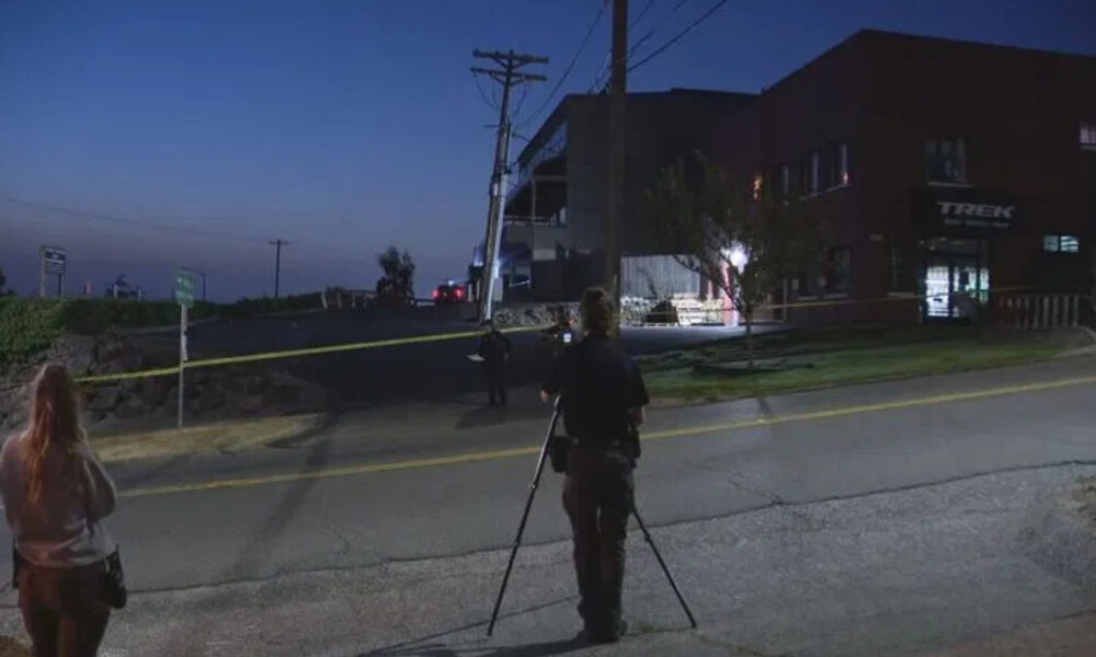 Nighttime street scene at a potential crime scene with yellow tape across the road, a person setting up a camera on a tripod, and investigators in the background near a brick building with a Trek sign.