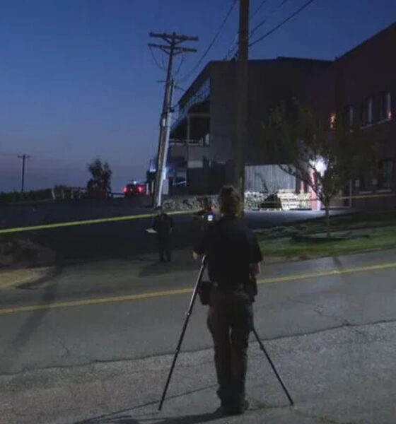 Nighttime street scene at a potential crime scene with yellow tape across the road, a person setting up a camera on a tripod, and investigators in the background near a brick building with a Trek sign.