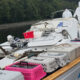 Work boat deck crowded with pet carriers, crates, and equipment; two people in white hazmat suits stand at the stern by the water