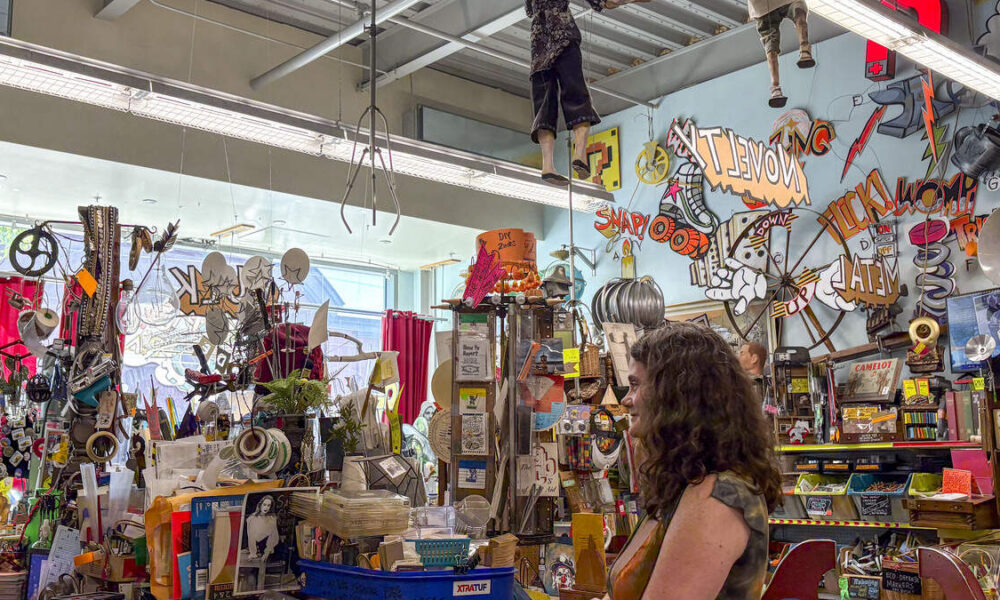 Cluttered art shop interior with a graffiti-covered wall, hanging props, and a woman browsing the displays.
