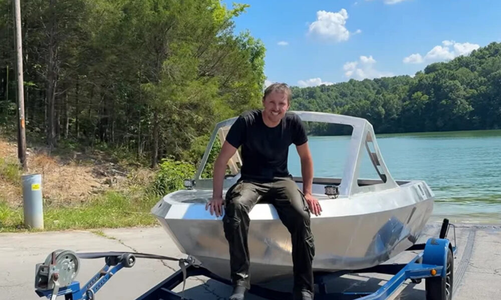 Man in a black t-shirt sits on a silver boat on a trailer by a lake, with trees in the background.
