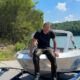 Man in a black t-shirt sits on a silver boat on a trailer by a lake, with trees in the background.