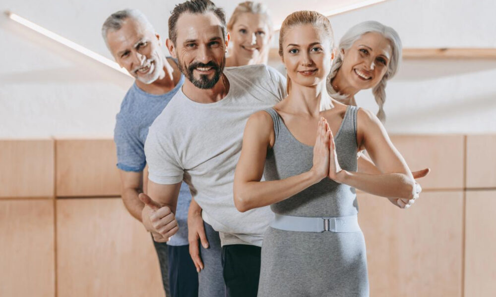 Group of adults in a fitness class standing in a line; front woman in gray with hands in prayer pose.