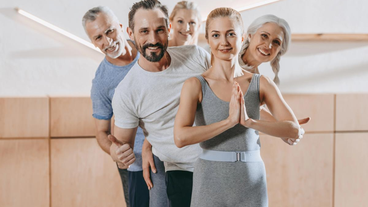 Group of adults in a fitness class standing in a line; front woman in gray with hands in prayer pose.