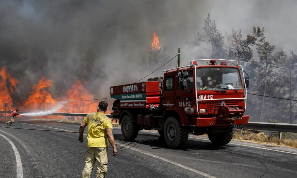 Firefighters battle a roadside wildfire as a red fire truck blocks the lane and flames rise behind a guardrail, smoke filling the air.