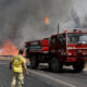 Firefighters battle a roadside wildfire as a red fire truck blocks the lane and flames rise behind a guardrail, smoke filling the air.