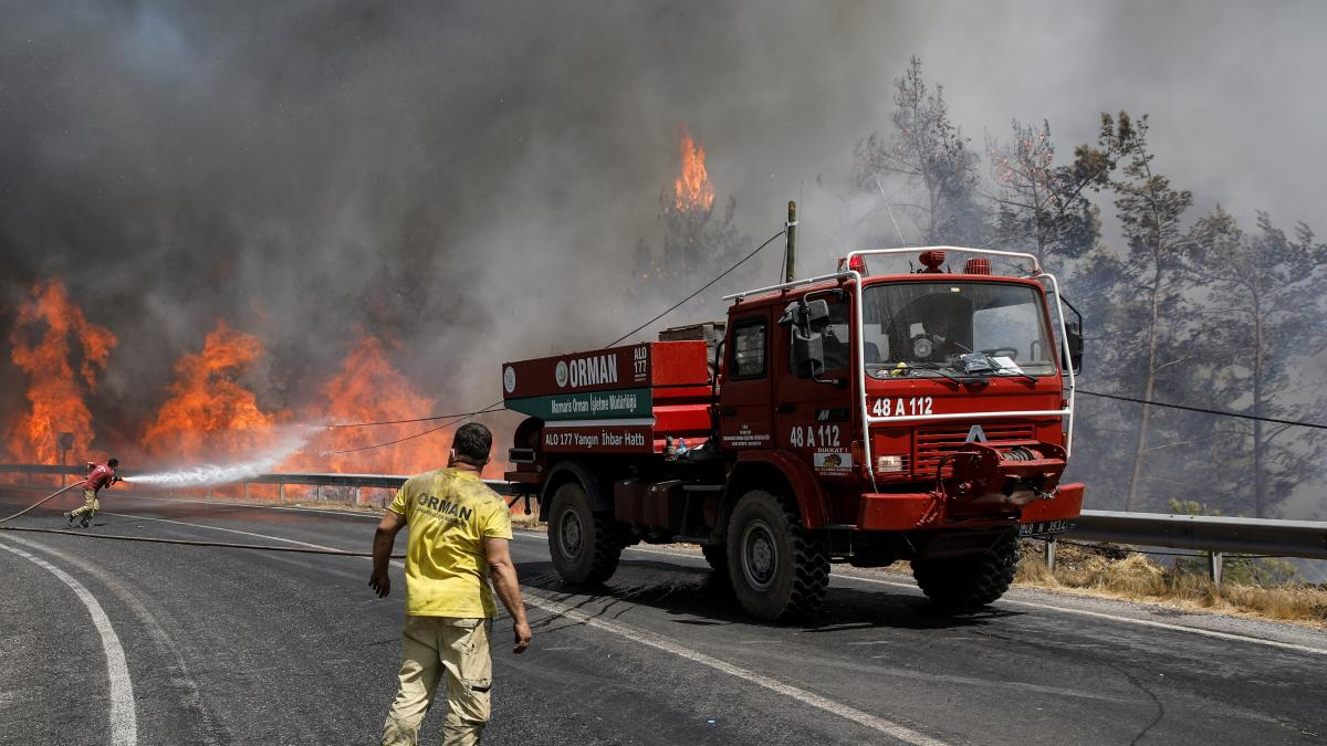 Firefighters battle a roadside wildfire as a red fire truck blocks the lane and flames rise behind a guardrail, smoke filling the air.