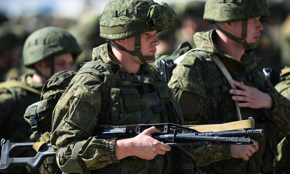 Soldier in camouflage gear and helmet holding an assault rifle, with fellow soldiers nearby.