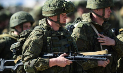 Soldier in camouflage gear and helmet holding an assault rifle, with fellow soldiers nearby.