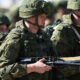 Soldier in camouflage gear and helmet holding an assault rifle, with fellow soldiers nearby.