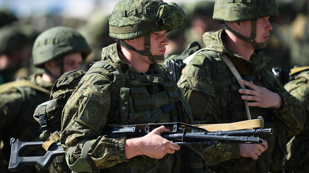 Soldier in camouflage gear and helmet holding an assault rifle, with fellow soldiers nearby.