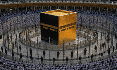 The Kaaba, a black cuboid with gold detailing, surrounded by pilgrims in white robes inside a large mosque courtyard.