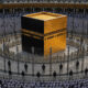 The Kaaba, a black cuboid with gold detailing, surrounded by pilgrims in white robes inside a large mosque courtyard.