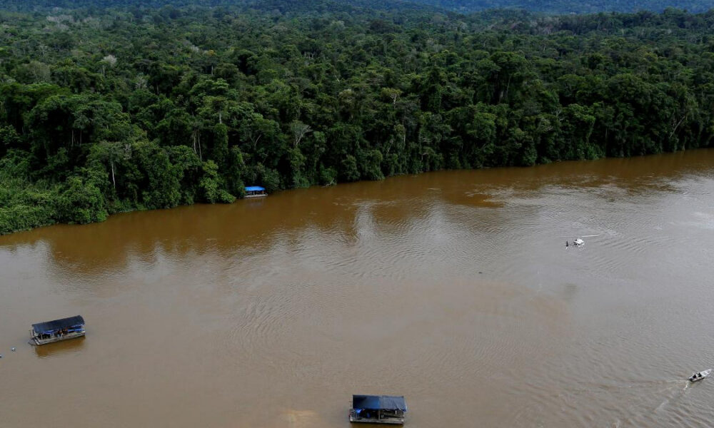 Aerial view of a wide muddy river flanked by dense green rainforest, with several boats under dark blue canopies on the water near the shore.