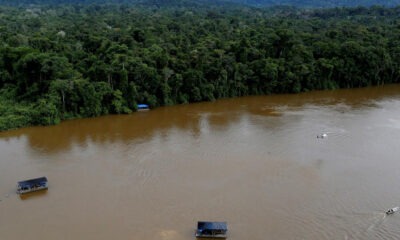 Aerial view of a wide muddy river flanked by dense green rainforest, with several boats under dark blue canopies on the water near the shore.
