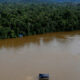 Aerial view of a wide muddy river flanked by dense green rainforest, with several boats under dark blue canopies on the water near the shore.