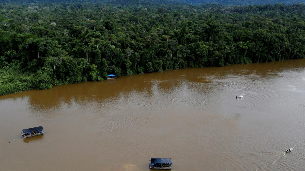Aerial view of a wide muddy river flanked by dense green rainforest, with several boats under dark blue canopies on the water near the shore.