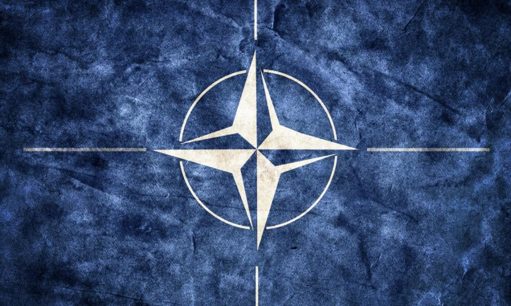NATO emblem: white compass rose inside a circle on a blue textured background.