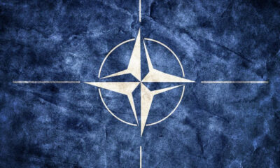 NATO emblem: white compass rose inside a circle on a blue textured background.