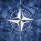 NATO emblem: white compass rose inside a circle on a blue textured background.