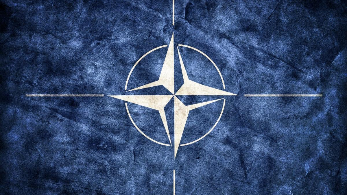 NATO emblem: white compass rose inside a circle on a blue textured background.