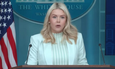 Blonde woman in a pale mint blazer speaks at a podium during a White House press briefing, with the American flag nearby.
