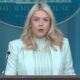 Blonde woman in a pale mint blazer speaks at a podium during a White House press briefing, with the American flag nearby.