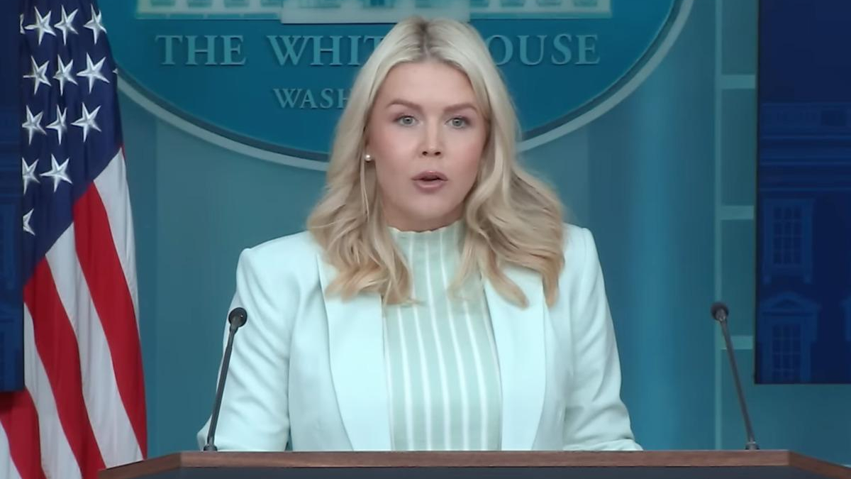 Blonde woman in a pale mint blazer speaks at a podium during a White House press briefing, with the American flag nearby.